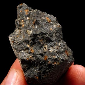 Könnte beinhalten: Ein raues, dunkelgraues Vulkangestein mit poröser, unebener Oberflächenstruktur und eingebetteten kleinen, metallisch orangefarbenen und weißen Mineraleinschlüssen, die über das gesamte gezackte, unregelmäßige Gesteinsfragment verteilt sind.