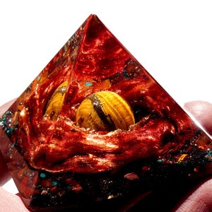 May include: A red, orange, and gold pyramid with a tiger's eye stone embedded inside. The pyramid is made of resin and has a sparkly, glittery finish.