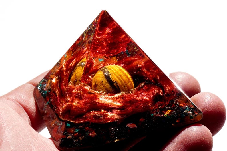 May include: A red, orange, and gold pyramid with a tiger's eye stone embedded inside. The pyramid is made of resin and has a sparkly, glittery finish.