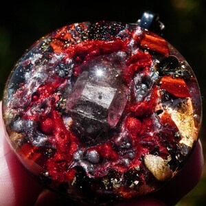 May include: A round, clear resin pendant with a red, gold, and silver design. A clear crystal is in the center of the pendant.