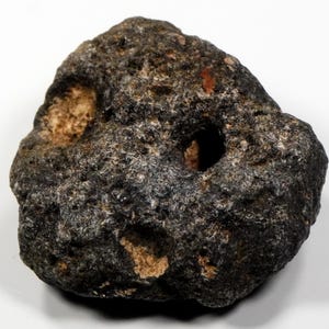 Könnte beinhalten: Ein dunkler, poröser Stein mit mehreren Löchern und hellbraunen Flecken. Der Stein hat eine raue, strukturierte Oberfläche und scheint eine natürliche geologische Formation zu sein. Das Bild ist vor einem weißen Hintergrund aufgenommen.