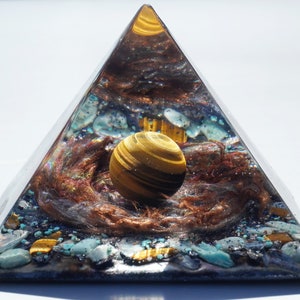 May include: A clear pyramid with a golden tiger's eye sphere inside. The pyramid is filled with various crystals and copper wire, creating a swirling, cosmic effect.