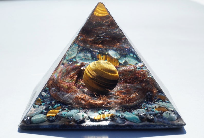 May include: A clear pyramid with a golden tiger's eye sphere inside. The pyramid is filled with various crystals and copper wire, creating a swirling, cosmic effect.
