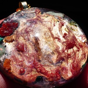 May include: A round, clear pendant with a gold-toned metallic finish. The pendant is filled with various colorful stones and glitter, including red, green, and gold.
