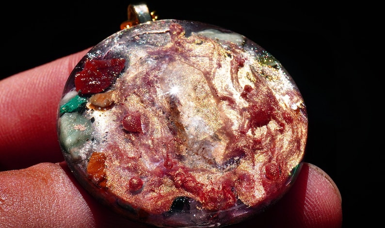 May include: A round, clear pendant with a gold-toned metallic finish. The pendant is filled with various colorful stones and glitter, including red, green, and gold.