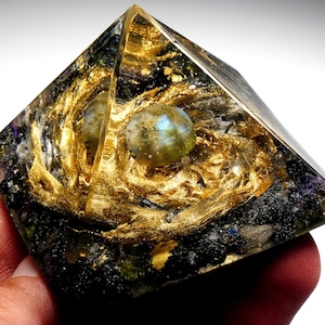 May include: A clear glass pyramid with gold leaf accents and two iridescent blue-green spheres inside. The pyramid is filled with black and white crystals.