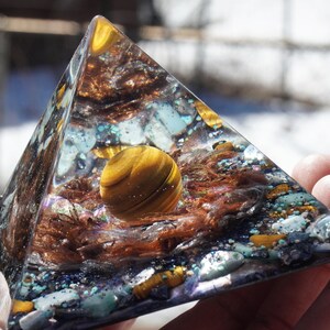 May include: A clear pyramid-shaped resin sculpture with a golden tiger's eye sphere inside. The sphere is surrounded by copper wire, iridescent glitter, and small pieces of turquoise and gold.