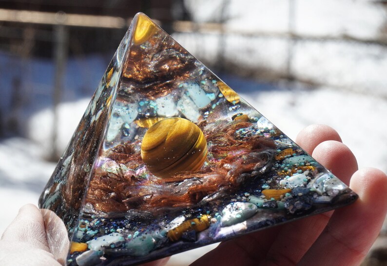 May include: A clear pyramid-shaped resin sculpture with a golden tiger's eye sphere inside. The sphere is surrounded by copper wire, iridescent glitter, and small pieces of turquoise and gold.