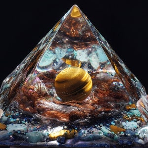 May include: A clear glass pyramid with a golden tiger's eye sphere inside. The pyramid is filled with various crystals and glitter, creating a mesmerizing effect.
