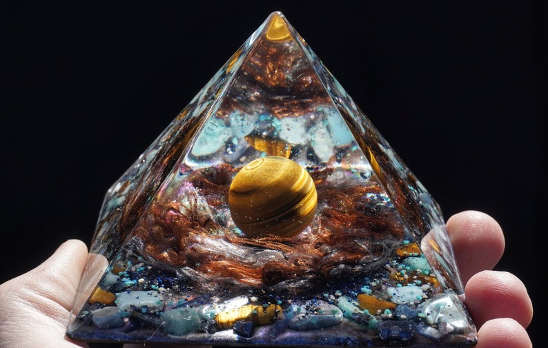 May include: A clear glass pyramid with a golden tiger's eye sphere inside. The pyramid is filled with various crystals and glitter, creating a mesmerizing effect.