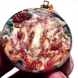 May include: A round, clear pendant with a gold chain. The pendant is filled with a swirling mixture of red, gold, and green glitter and small pieces of various colored stones.