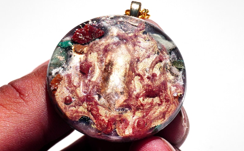 May include: A round, clear pendant with a gold chain. The pendant is filled with a swirling mixture of red, gold, and green glitter and small pieces of various colored stones.
