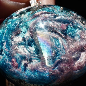May include: A round, iridescent, blue and pink resin pendant with a silver bail. The pendant has a swirling, marbled pattern and a clear, iridescent crystal in the center.