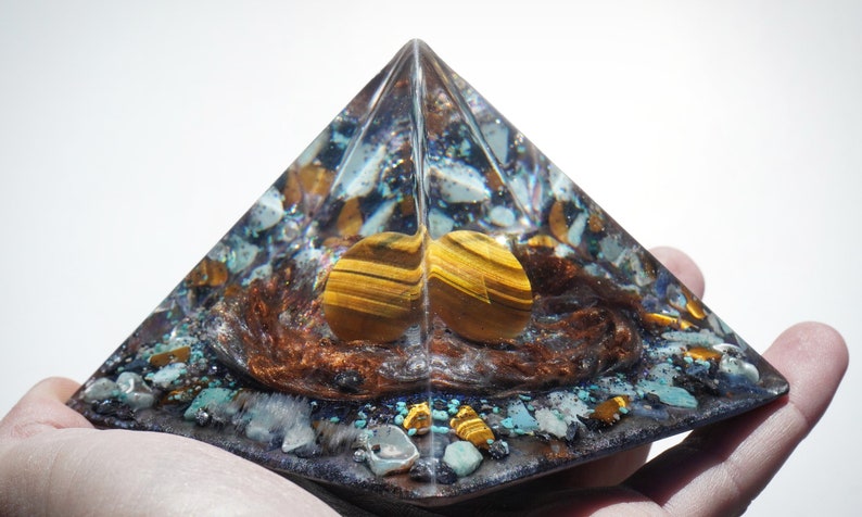 May include: A clear pyramid-shaped crystal with a brown, gold, and blue interior. The crystal contains small pieces of various stones, including tiger's eye, turquoise, and copper.
