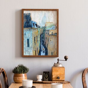 May include: A framed painting of a city street scene with a church dome in the distance. The painting is done in a watercolor style with muted colors of blue, gray, and yellow. The frame is a simple brown wood frame.