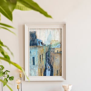 May include: A framed painting of a city street scene with buildings and a dome in the distance. The painting is in a white frame and is hanging on a white wall. There is a plant in the foreground.