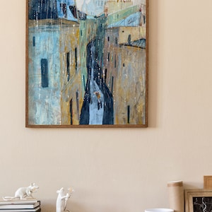 May include: A framed painting of a city street scene with buildings and a church in the background. The painting is done in a watercolor style with a muted color palette of blues, yellows, and browns. The painting is framed in a brown wood frame.