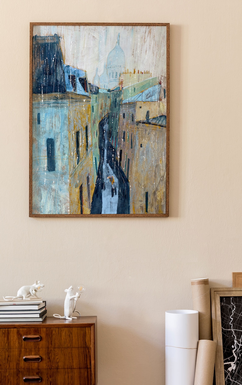 May include: A framed painting of a city street scene with buildings and a church in the background. The painting is done in a watercolor style with a muted color palette of blues, yellows, and browns. The painting is framed in a brown wood frame.