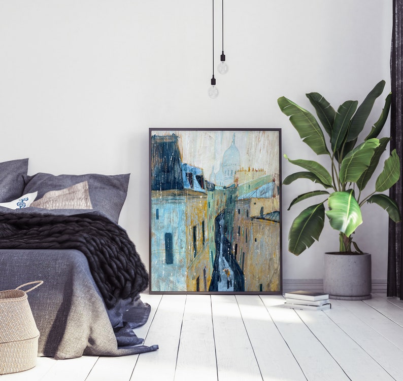 May include: A painting of a Parisian street scene with a view of the Sacr&eacute;-C&oelig;ur Basilica in the distance. The painting is in a black frame and is hanging on a white wall.