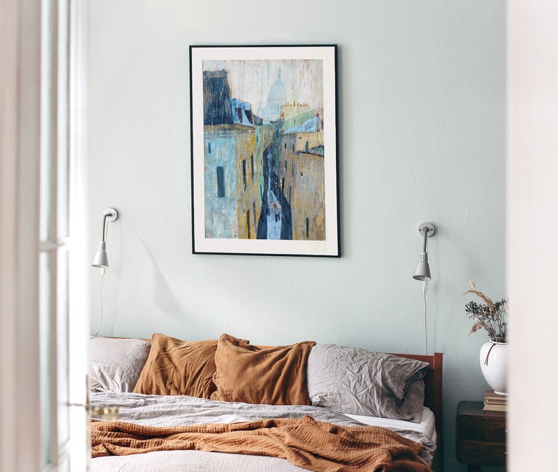 May include: A bedroom with a bed with brown and gray bedding and a framed painting of a city skyline with a church in the background. The painting is hanging on a light blue wall. There are two wall sconces on either side of the painting.