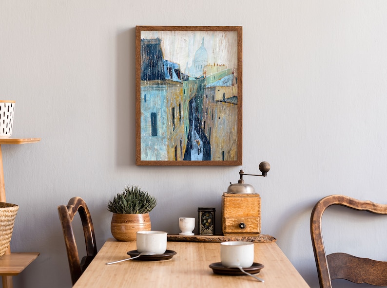 May include: A framed painting of a city street scene with a church dome in the distance. The painting is done in a watercolor style with muted colors of blue, gray, and yellow. The frame is a simple brown wood frame.