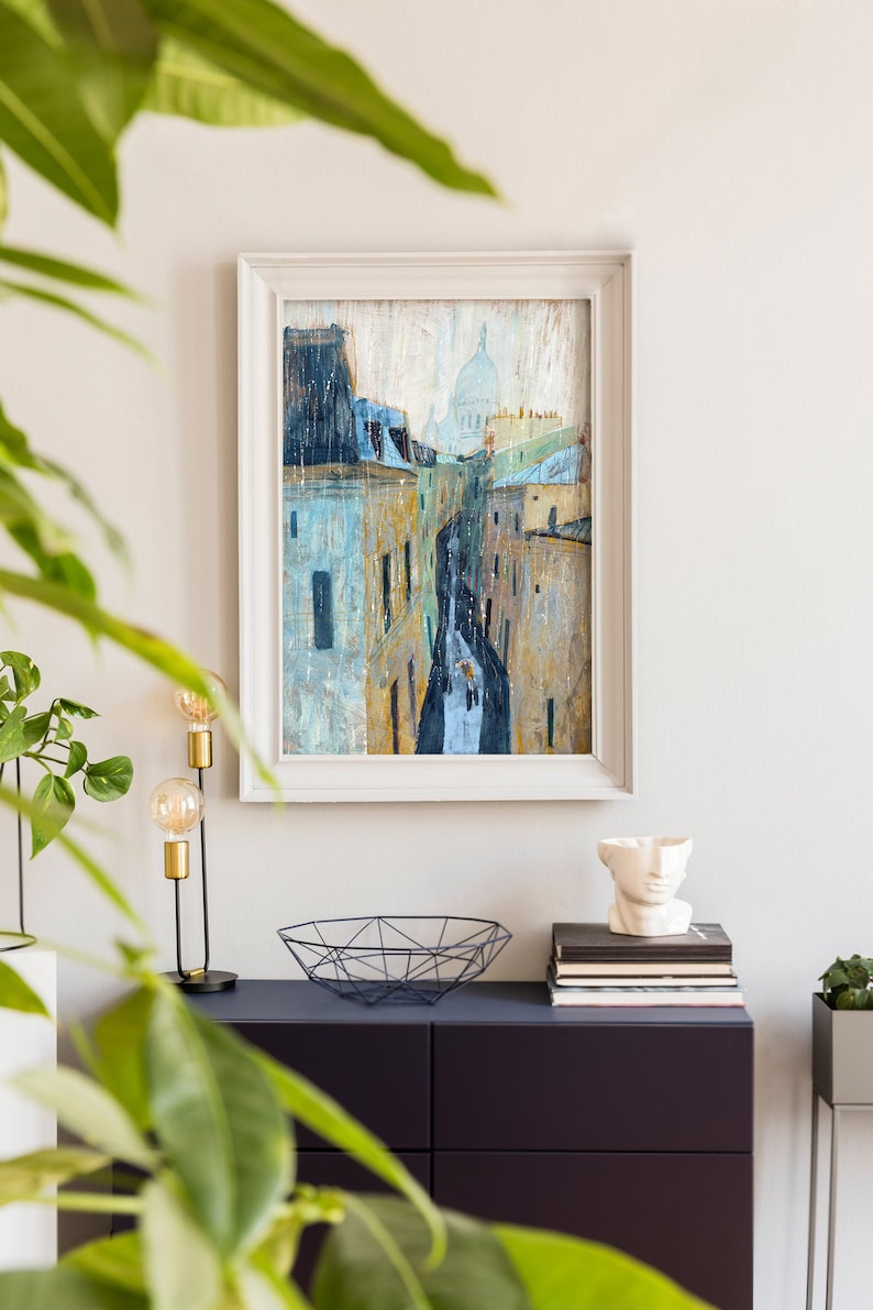 May include: A framed painting of a city street scene with buildings and a dome in the distance. The painting is in a white frame and is hanging on a white wall. There is a plant in the foreground.