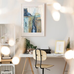 May include: A white desk with a wooden chair, a vintage typewriter, a rotary phone, and a framed painting of a city street with buildings. The desk is set against a white wall with a white shelf unit behind it. The room is decorated with string lights.