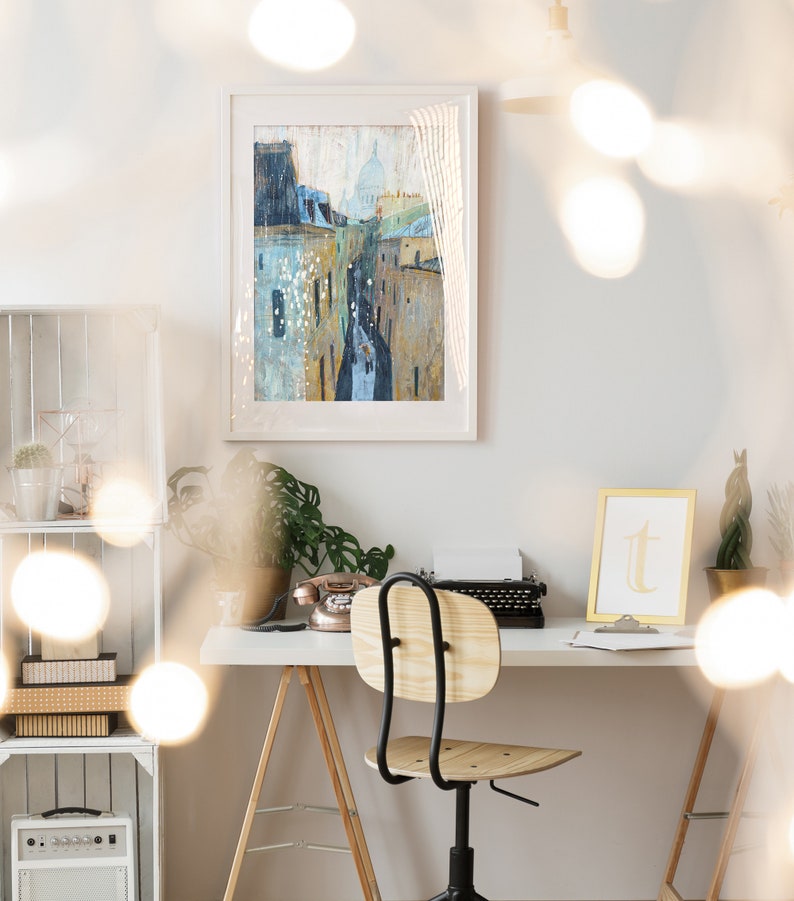 May include: A white desk with a wooden chair, a vintage typewriter, a rotary phone, and a framed painting of a city street with buildings. The desk is set against a white wall with a white shelf unit behind it. The room is decorated with string lights.