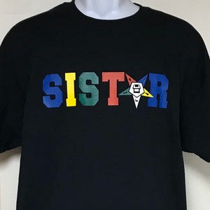 May include: Black t-shirt with the word "SISTAR" in colorful block letters and a five-pointed star with a Masonic symbol in the center.