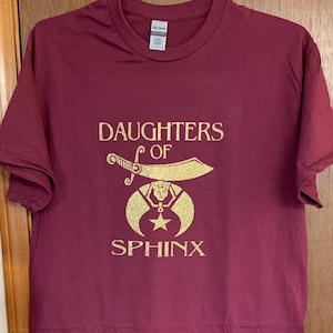 May include: A maroon t-shirt with a gold glitter design that says "Daughters of Sphinx". The design features a crescent moon, a star, and a sword.