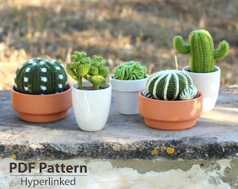 May include: A collection of five crocheted cactus plants in various styles. The cacti are in small terracotta and white ceramic pots. The cacti are green with white accents. The image includes the text "PDF Pattern Hyperlinked".