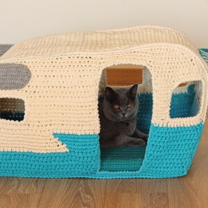 May include: A crocheted cat bed shaped like a vintage camper. The camper is white with a blue bottom and a gray awning. A gray cat is sitting inside the camper.