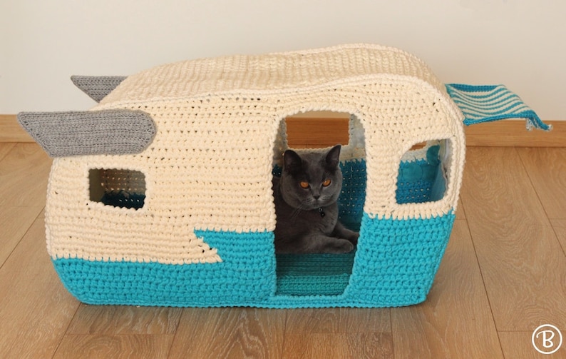 May include: A crocheted cat bed shaped like a vintage camper. The camper is white with a blue bottom and a gray awning. A gray cat is sitting inside the camper.