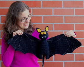 May include: A person holding a black crocheted bat with yellow eyes. The bat has a long tail and is made of yarn.