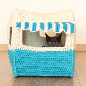 May include: A crocheted cat bed shaped like a small house with a blue and white striped awning. A gray cat is looking out of the window.