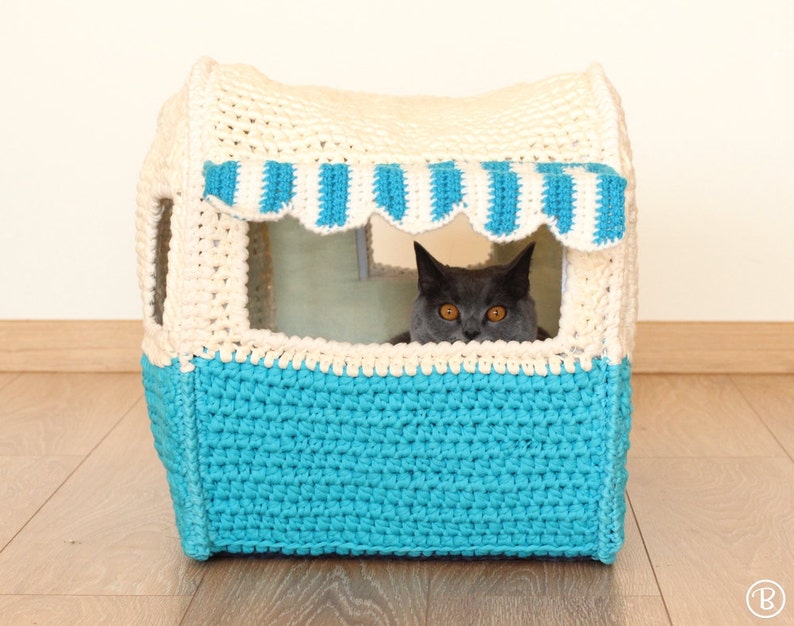 May include: A crocheted cat bed shaped like a small house with a blue and white striped awning. A gray cat is looking out of the window.