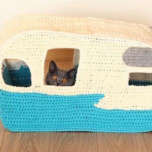 May include: A crocheted cat bed shaped like a vintage camper. The camper is white with a blue bottom and a gray awning. A cat is peeking out of the window.