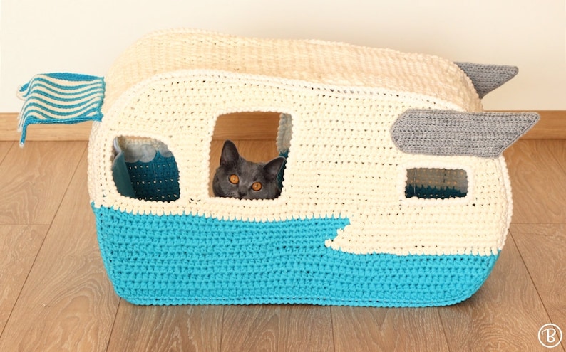 May include: A crocheted cat bed shaped like a vintage camper. The camper is white with a blue bottom and a gray awning. A cat is peeking out of the window.