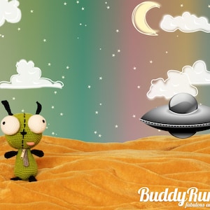 May include: A green crocheted alien amigurumi with large white eyes and black antennae stands on a textured orange surface. A gray UFO hovers in the background, with a rainbow, moon, and clouds in a starry sky. The image has the text "BuddyRumi.com".