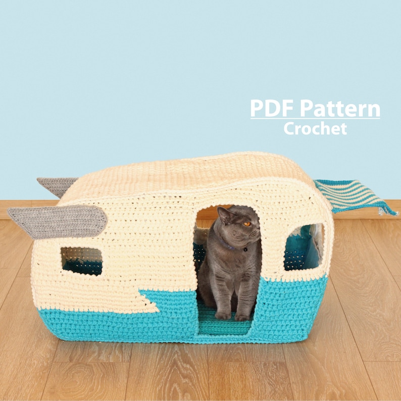 May include: Crochet pattern for a cat bed shaped like a camper van. The bed is white and blue with a gray awning. A gray cat is sitting inside the bed.