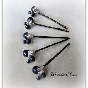 May include: Six black hair pins with a cluster of three black pearls and a rhinestone on each pin. The pins are arranged in a fan shape on a white surface.