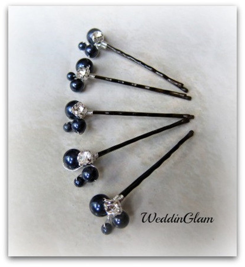 May include: Six black hair pins with a cluster of three black pearls and a rhinestone on each pin. The pins are arranged in a fan shape on a white surface.