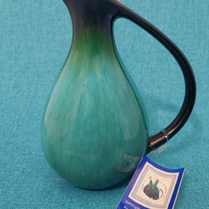 May include: A teal-colored ceramic pitcher with a black handle and rim. The pitcher has a teardrop shape and a glossy finish. A tag with text is attached to the pitcher.