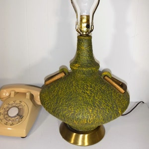 May include: A green and yellow ceramic table lamp with a brass base and wooden accents. The lamp has a textured surface and a round, wide base.