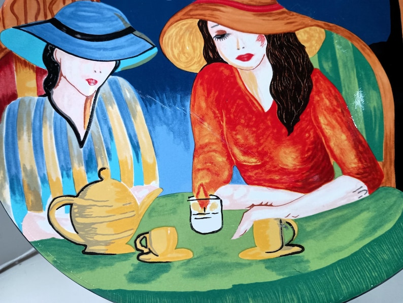 Furio Café plates displayed together: a large oval dinner plate featuring two women seated at a café table with a yellow teapot and cups, with bold, colorful artwork