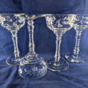 May include: Four clear, etched glass champagne coupe glasses. Each glass features a floral design around the rim and on the base. The stems are intricately designed. The glasses are arranged on a blue background.