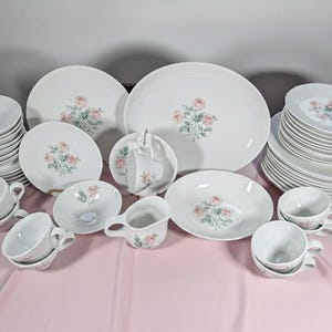 May include: A white porcelain dinnerware set featuring a floral rose design. The set includes plates, bowls, cups, and saucers. The roses are pink and green. The set is arranged on a pink tablecloth.