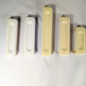 May include: Five ceramic door pulls in various sizes. Two are white, two are cream, and one is a smaller cream color. All have a rectangular shape with a recessed center.