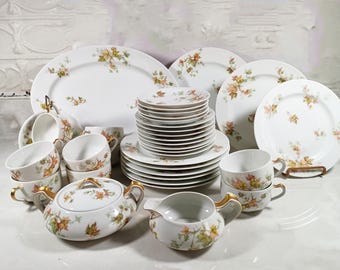 May include: A set of white porcelain dinnerware with a delicate floral pattern featuring autumn leaves in shades of orange, brown, and yellow. The set includes plates, bowls, cups, and a creamer.