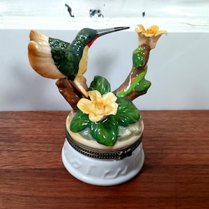 May include: A ceramic hummingbird figurine perched on a branch with yellow flowers and green leaves. The bird has green, tan, and red coloring. The figurine sits atop a white, round, lidded box with a decorative metal band.